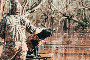 Man in camouflage patting black lab hunting dog on platform in flooded woodland