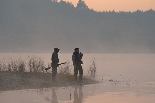Two hunters with shotguns stand on a misty lakeshore at dawn, silhouetted against foggy forest backdrop