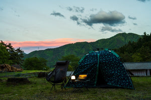 Camping tent, folding chair and laptop on mountain campsite at sunset with pink sky and green forested hills
